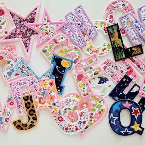 May include: A collection of fabric letters and a star, each with a different floral pattern. The letters are in various colours, including pink, purple, blue, and green. The star is pink with a white floral pattern.