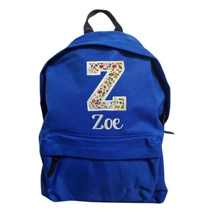 May include: A royal blue backpack featuring a large letter Z patch with a colorful floral pattern. Below the letter, the name Zoe is embroidered in white script. The backpack includes a top handle and a front zippered pocket for storage.