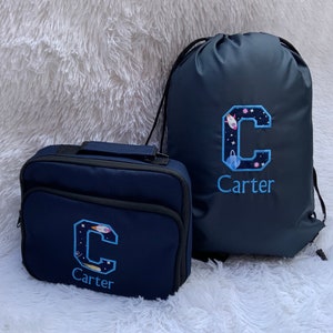 May include: A navy blue lunch bag and drawstring bag with a space-themed embroidered letter C and the name Carter.