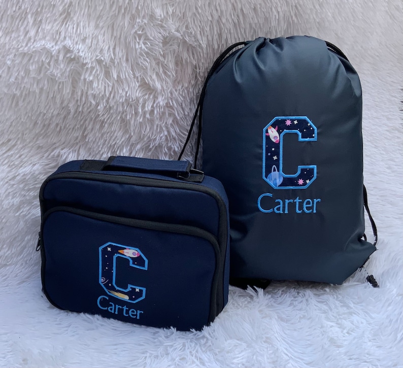 May include: A navy blue lunch bag and drawstring bag with a space-themed embroidered letter C and the name Carter.