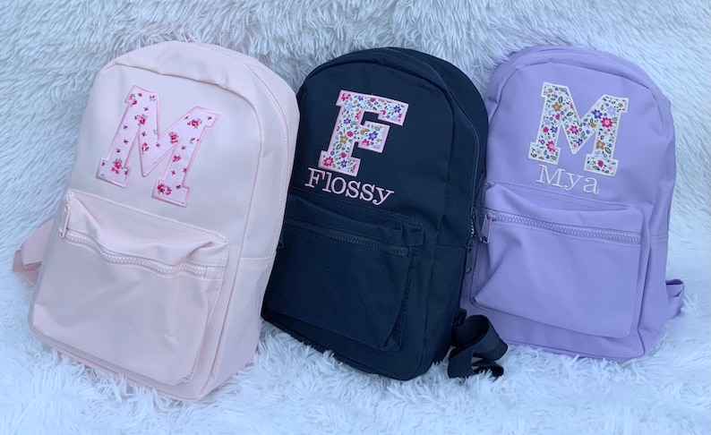 May include: Three backpacks in light pink, black, and light purple. The pink backpack features a floral letter M. The black backpack has a floral letter F and the name Flossy. The purple backpack has a floral letter M and the name Mya.