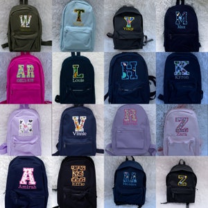 May include: A collection of sixteen backpacks in various solid colors, each featuring a large embroidered initial letter with a patterned fill and a personalized name stitched underneath. The backpacks include front zippered pockets and shoulder straps.