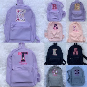 May include: A collection of small backpacks in various solid colors including lavender, pink, black, and gray. Each backpack features a front zippered pocket and a large, personalized embroidered letter patch with unique patterns and names stitched below.