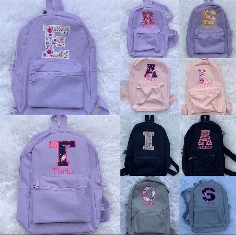 May include: A collection of small backpacks in various solid colors including lavender, pink, black, and gray. Each backpack features a front zippered pocket and a large, personalized embroidered letter patch with unique patterns and names stitched below.
