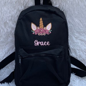 May include: Black canvas backpack with a unicorn design embroidered on the front. The unicorn has a gold horn, pink flowers, and pink eyelashes. The name "Grace" is embroidered below the unicorn.