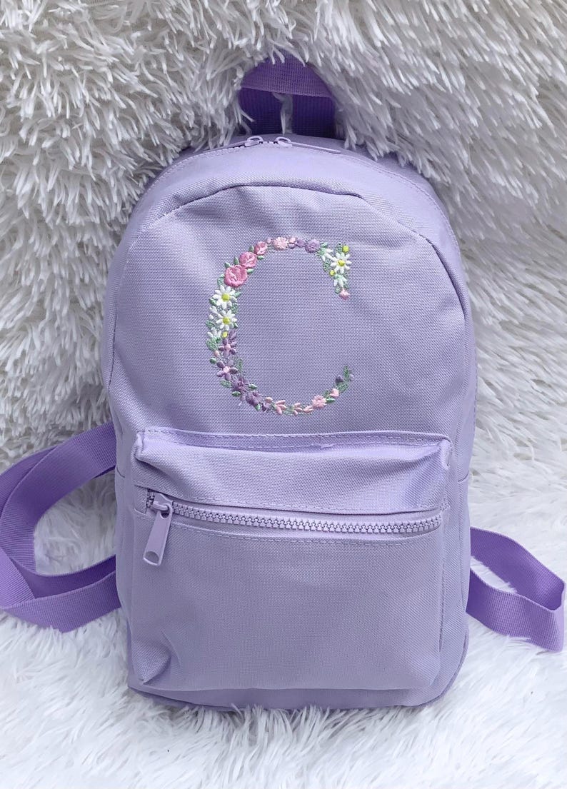 May include: A lavender purple mini backpack with a floral embroidered letter C on the front. The backpack has a zippered pocket on the front and adjustable straps.