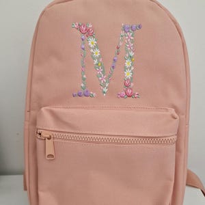 May include: A pink canvas backpack with a floral embroidered letter "M" on the front. The backpack has a zippered front pocket.