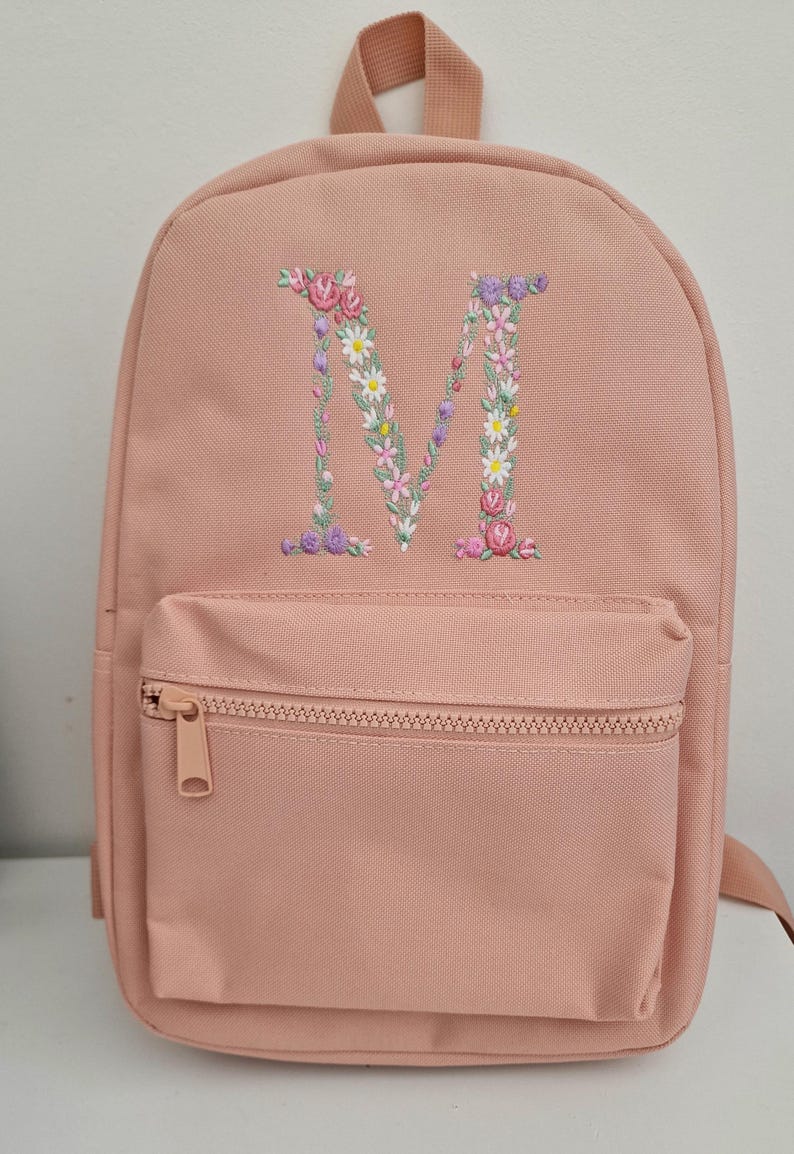 May include: A pink canvas backpack with a floral embroidered letter "M" on the front. The backpack has a zippered front pocket.
