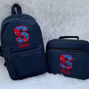 May include: A blue backpack and lunch bag set with a red embroidered letter "S" and the name "Shay" on both. The letter "S" is decorated with a Spiderman graphic.