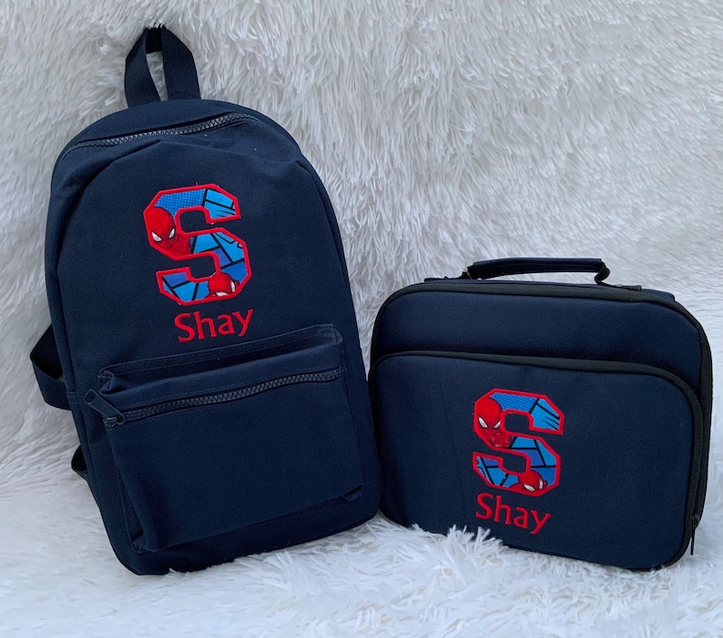 May include: A blue backpack and lunch bag set with a red embroidered letter "S" and the name "Shay" on both. The letter "S" is decorated with a Spiderman graphic.