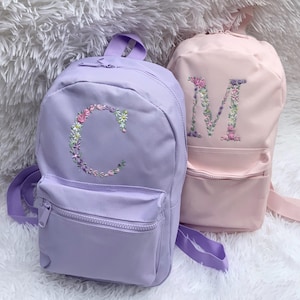 May include: Two purple and pink backpacks with floral embroidered letters C and M. The backpacks have a front pocket with a zipper.