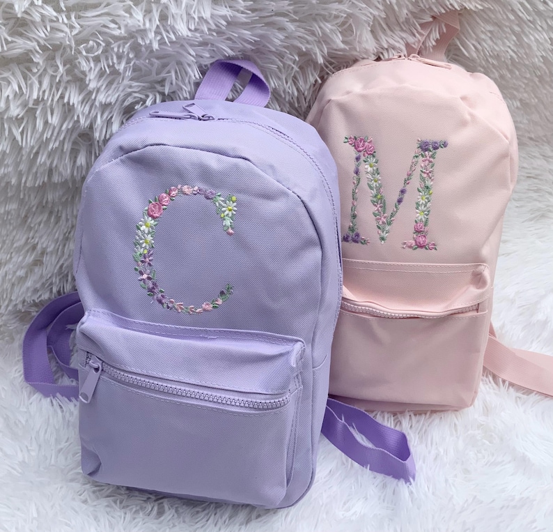 May include: Two purple and pink backpacks with floral embroidered letters C and M. The backpacks have a front pocket with a zipper.
