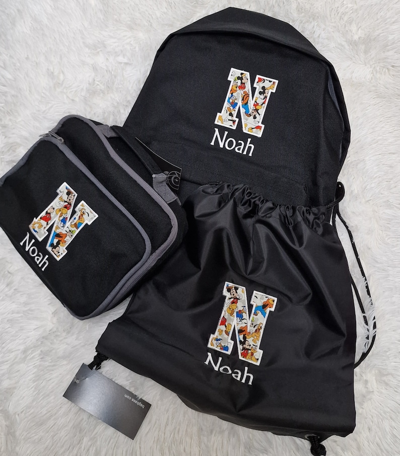 May include: A black backpack, lunch bag, and drawstring bag set with a Mickey Mouse character print. Each bag has a white letter 'N' with the name 'Noah' embroidered below.