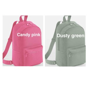 May include: Two backpacks shown side by side. The left backpack is candy pink and the right backpack is dusty green. Each bag features a main zippered compartment, a smaller front zippered pocket, adjustable shoulder straps, and a top handle.