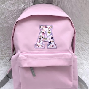 Puede incluir: Una mochila rosa claro con un bolsillo frontal y correas grises. La mochila presenta una gran letra "A" bordada con flores en tonos rosa, morado y blanco. La bolsa tiene un asa superior y un bolsillo frontal con cremallera.