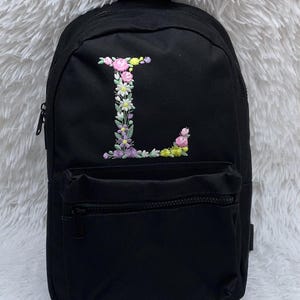 May include: Black canvas backpack with a floral embroidered letter L on the front. The backpack has a zippered pocket on the front and a zippered main compartment.