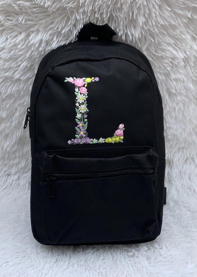 May include: Black canvas backpack with a floral embroidered letter L on the front. The backpack has a zippered pocket on the front and a zippered main compartment.