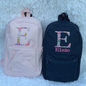 May include: Two personalized backpacks with floral embroidery. The pink backpack has a floral letter E. The black backpack has a floral letter E and the name Elsie embroidered below it.