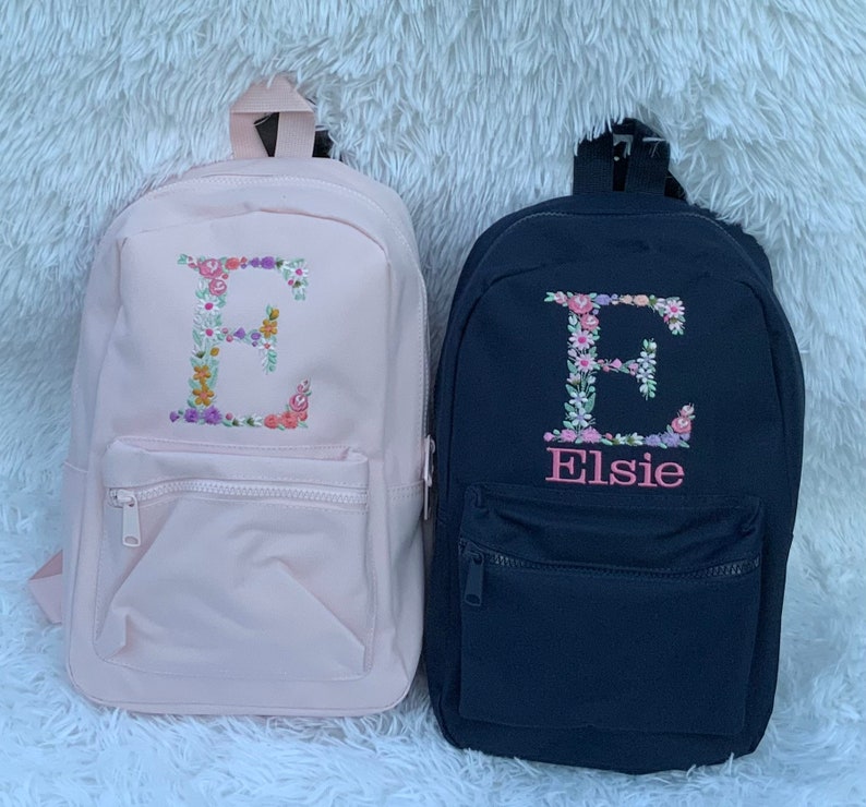 May include: Two personalized backpacks with floral embroidery. The pink backpack has a floral letter E. The black backpack has a floral letter E and the name Elsie embroidered below it.