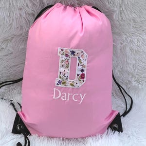 May include: Pink drawstring bag with a floral letter D and the name Darcy embroidered on the front.