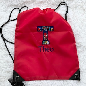 K&ouml;nnte beinhalten: Ein roter Kordelzugrucksack mit einem gro&szlig;en Buchstaben T, der ein Comic-Muster mit einer Spider-Man-Grafik aufweist. Unter dem Buchstaben ist der Name Theo in blauem Garn gestickt. Der Rucksack hat schwarze Kordeltr&auml;ger und schwarze Eckverst&auml;rkungen.