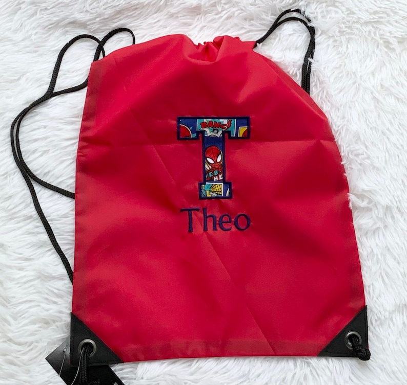K&ouml;nnte beinhalten: Ein roter Kordelzugrucksack mit einem gro&szlig;en Buchstaben T, der ein Comic-Muster mit einer Spider-Man-Grafik aufweist. Unter dem Buchstaben ist der Name Theo in blauem Garn gestickt. Der Rucksack hat schwarze Kordeltr&auml;ger und schwarze Eckverst&auml;rkungen.
