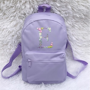 May include: A lavender purple backpack with a front zippered pocket. The backpack has a floral embroidered letter "E" on the front.