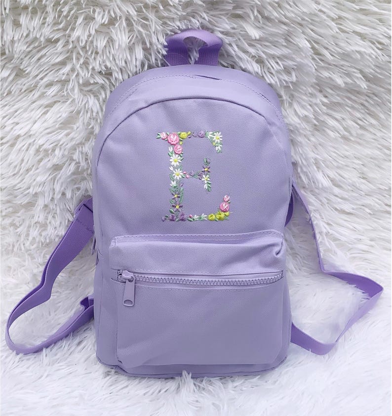 May include: A lavender purple backpack with a front zippered pocket. The backpack has a floral embroidered letter "E" on the front.