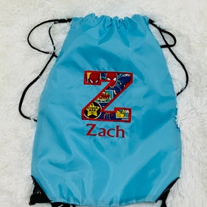 K&ouml;nnte beinhalten: Ein hellblauer Kordelzugrucksack mit einem gro&szlig;en Buchstaben Z, der in einem Comic-Druck mit Spider-Man-Motiven appliziert ist. Der Name Zach ist unter dem Buchstaben in rotem Garn gestickt. Die Tasche hat schwarze Kordeltr&auml;ger und eine glatte Oberfl&auml;che.