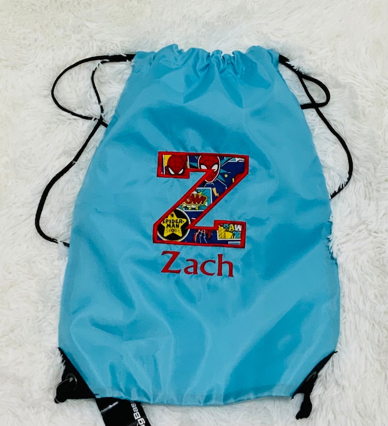 K&ouml;nnte beinhalten: Ein hellblauer Kordelzugrucksack mit einem gro&szlig;en Buchstaben Z, der in einem Comic-Druck mit Spider-Man-Motiven appliziert ist. Der Name Zach ist unter dem Buchstaben in rotem Garn gestickt. Die Tasche hat schwarze Kordeltr&auml;ger und eine glatte Oberfl&auml;che.