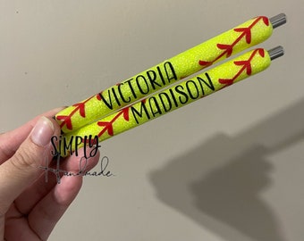 May include: Two yellow glitter pens with red stitching details, resembling softballs. The pens are personalized with the names "VICTORIA" and "MADISON" in black lettering. The pens are held in a hand.