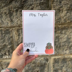 May include: A white notepad with a red border, featuring the text "Mrs. Taylor" at the top. The notepad has a decorative pumpkin sticker and the words "SIMPLY handmade" in cursive.