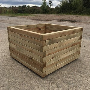 May include: A square wooden planter box made of natural wood planks. The planter is empty and sits on a gravel surface.