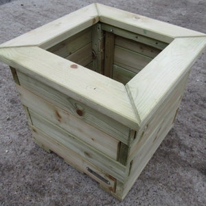 May include: A square wooden planter with a light brown finish. The planter has a raised lip around the top and is made of multiple pieces of wood.