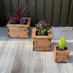 May include: Three wooden planter boxes in different sizes, with various plants growing inside. The largest planter is brown and has a dark red plant with pink flowers. The middle planter is brown and has a red plant with small white flowers. The smallest planter is brown and has a small green plant.