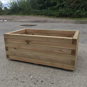 May include: A rectangular wooden planter box with a natural wood finish. The planter is made of horizontal planks and has a simple, rustic design.