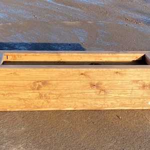 May include: A rectangular wooden planter box with a natural wood finish. The planter is made of horizontal planks of wood and has a simple design.