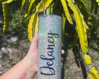 Può includere: Un bicchiere trasparente e iridescente con il nome "Delaney" scritto in nero in corsivo sul lato. Il bicchiere è ricoperto di strass ed è tenuto da una mano con un tatuaggio di scorpione.
