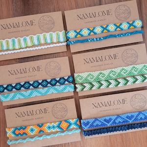 May include: Six pairs of colourful friendship bracelets on brown card stock with the text "NAMALOMÉ HANDMADE JEWELRY" on each card.