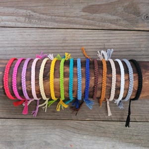 May include: A collection of fourteen woven cord bracelets in a spectrum of colours including red, pink, lavender, light pink, cream, yellow, lime green, turquoise, blue, dark blue, brown, orange, beige, white, grey, and black with small tassels.