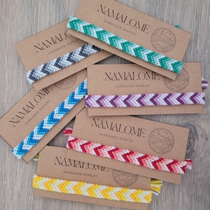 Peut inclure: Six bracelets d'amitié colorés avec des motifs en chevron sur des cartes brunes avec le texte "NAMALOMÉ HANDMADE JEWELRY".
