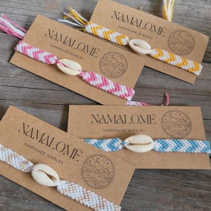 May include: Four woven friendship bracelets featuring a chevron pattern in pink, yellow, light blue, and beige, each centered with a white cowrie shell. The bracelets are attached to brown cardboard display cards labeled NAMALOME.