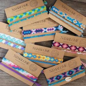 May include: A selection of handmade friendship bracelets in diverse colours and patterns, presented on brown cardboard tags. The bracelets showcase complex woven designs in shades of blue, purple, pink, and yellow. Each tag is labelled "NAMALOME HANDMADE JEWELRY".