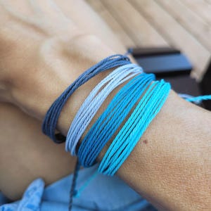 Peut inclure: Plusieurs bracelets en ficelle bleus et turquoise enroulés autour d'un poignet. Les bracelets varient en nuances, du bleu foncé au turquoise clair. Les ficelles sont étroitement tissées, créant un aspect texturé. Accessoire simple et minimaliste.
