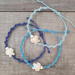Peut inclure: Trois bracelets faits à la main en nuances de bleu, avec des breloques tortues. Les bracelets sont faits de cordon torsadé et ont des fermetures réglables. Les breloques tortues sont blanc cassé et ont des motifs de coquille détaillés.