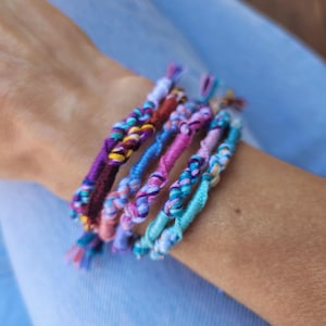 May include: A selection of brightly coloured braided friendship bracelets. The bracelets display a range of hues, including purple, blue, pink, and teal. Each bracelet is intricately woven with a unique pattern, demonstrating the craftsmanship.