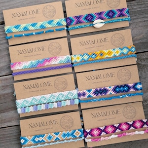 May include: Eight pairs of colourful friendship bracelets on brown card stock with the text "NAMALOME HANDMADE JEWELRY" printed on each card.