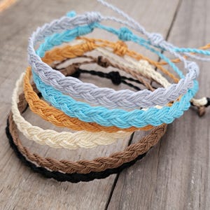 Peut inclure: Plusieurs bracelets en corde tressée de différentes couleurs, dont bleu clair, beige, marron et noir. Les bracelets sont noués pour être ajustables. Ils sont présentés sur une surface en bois vieilli.