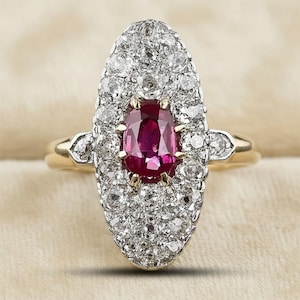 May include: A vintage-style ring featuring a central oval-shaped ruby surrounded by numerous small diamonds. The ring has a gold band and a halo of diamonds around the ruby, creating a sparkling effect. The ruby is a deep red color.
