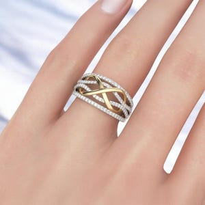 K&ouml;nnte beinhalten: Ein gold-silberner Ring mit einem Unendlichkeits-Symbol-Design. Der Ring hat ein goldenes Unendlichkeits-Symbol in der Mitte, umgeben von silbernen B&auml;ndern, die mit kleinen, klaren Edelsteinen besetzt sind. Der Ring befindet sich an einem Finger.